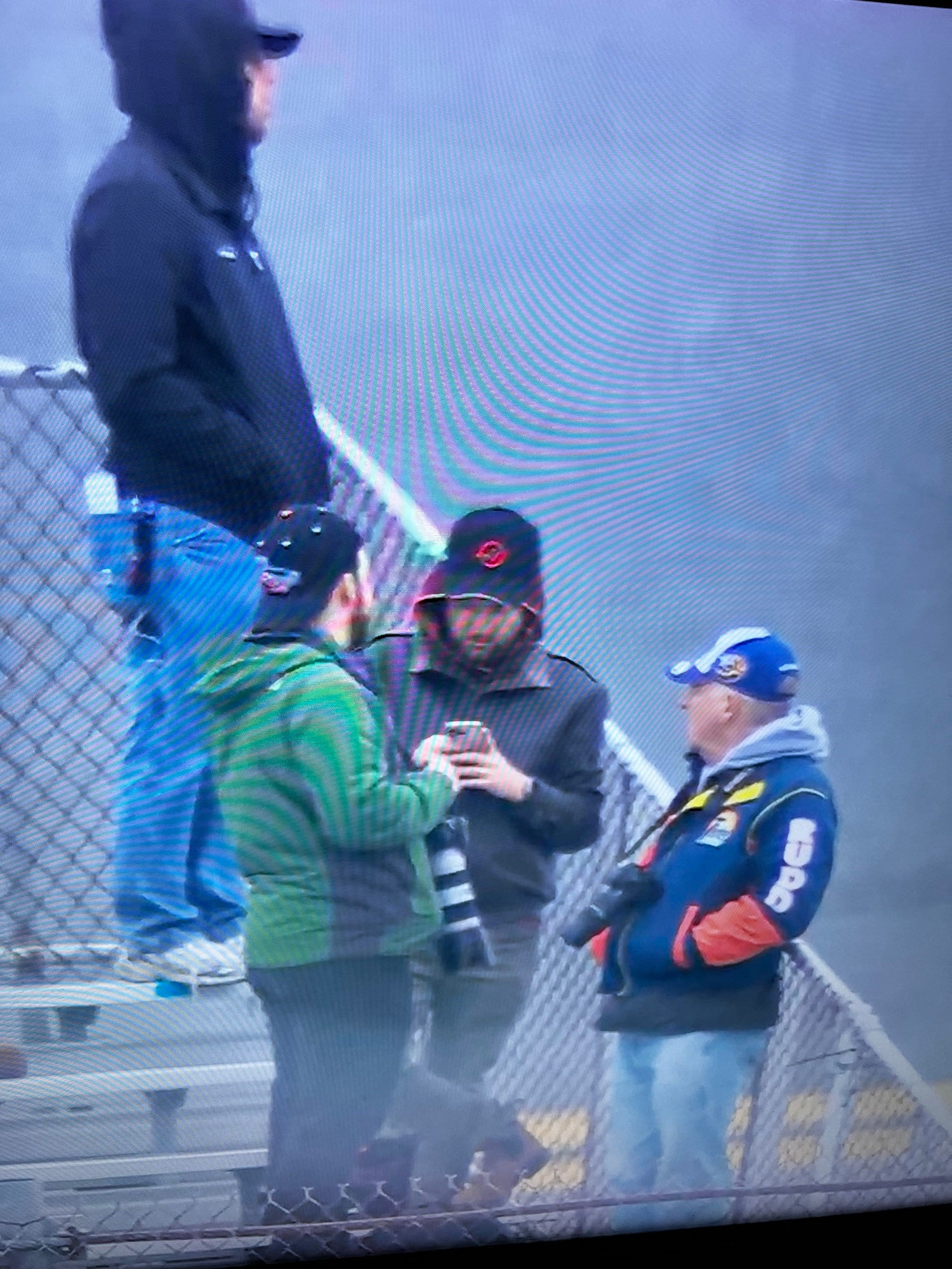 A photograph of a TV screen showing people in rain gear in bleachers looking at screens