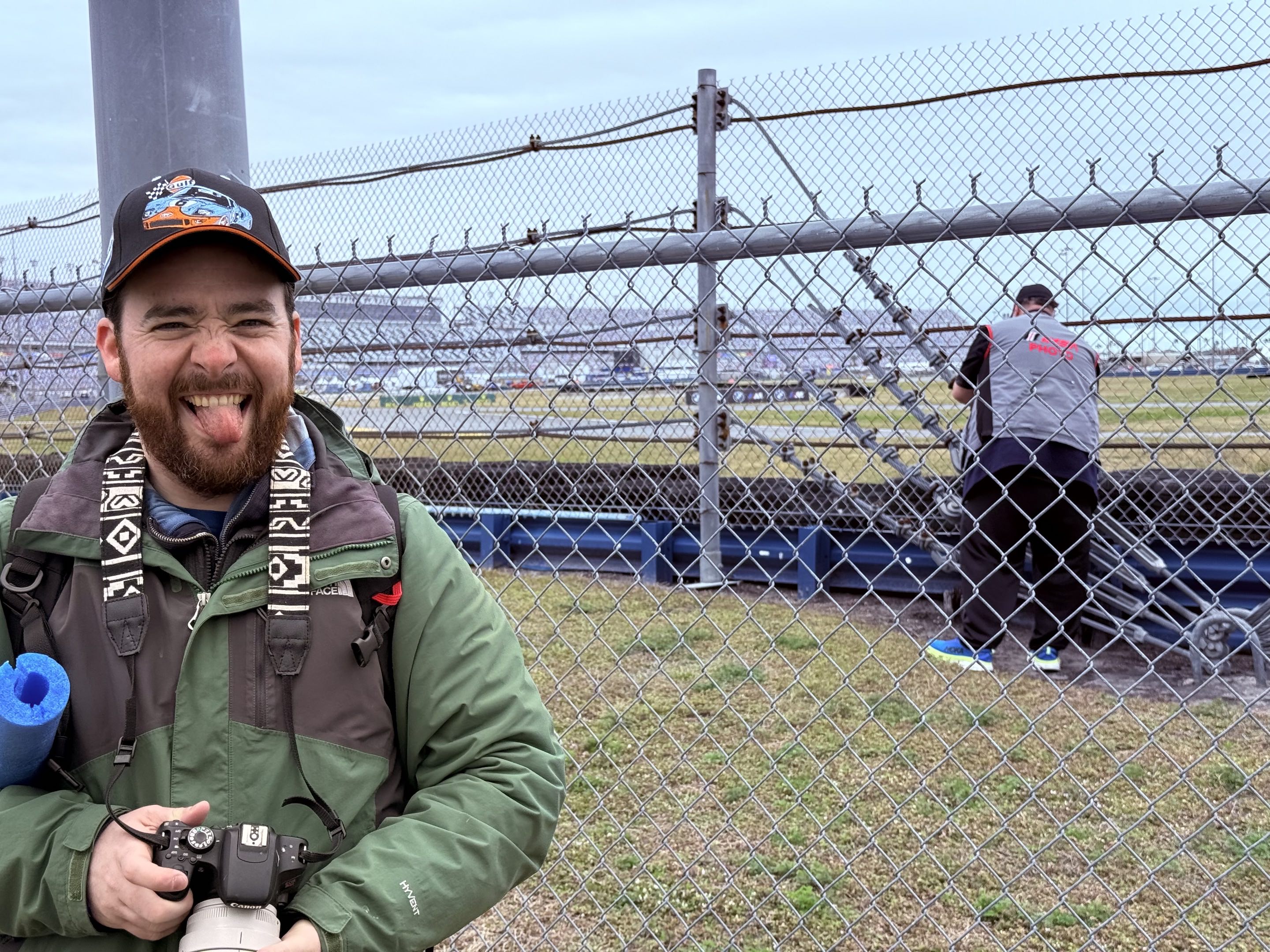 A thirtysomething man makes a very happy face while a large photojournalist aims at the race track behind him