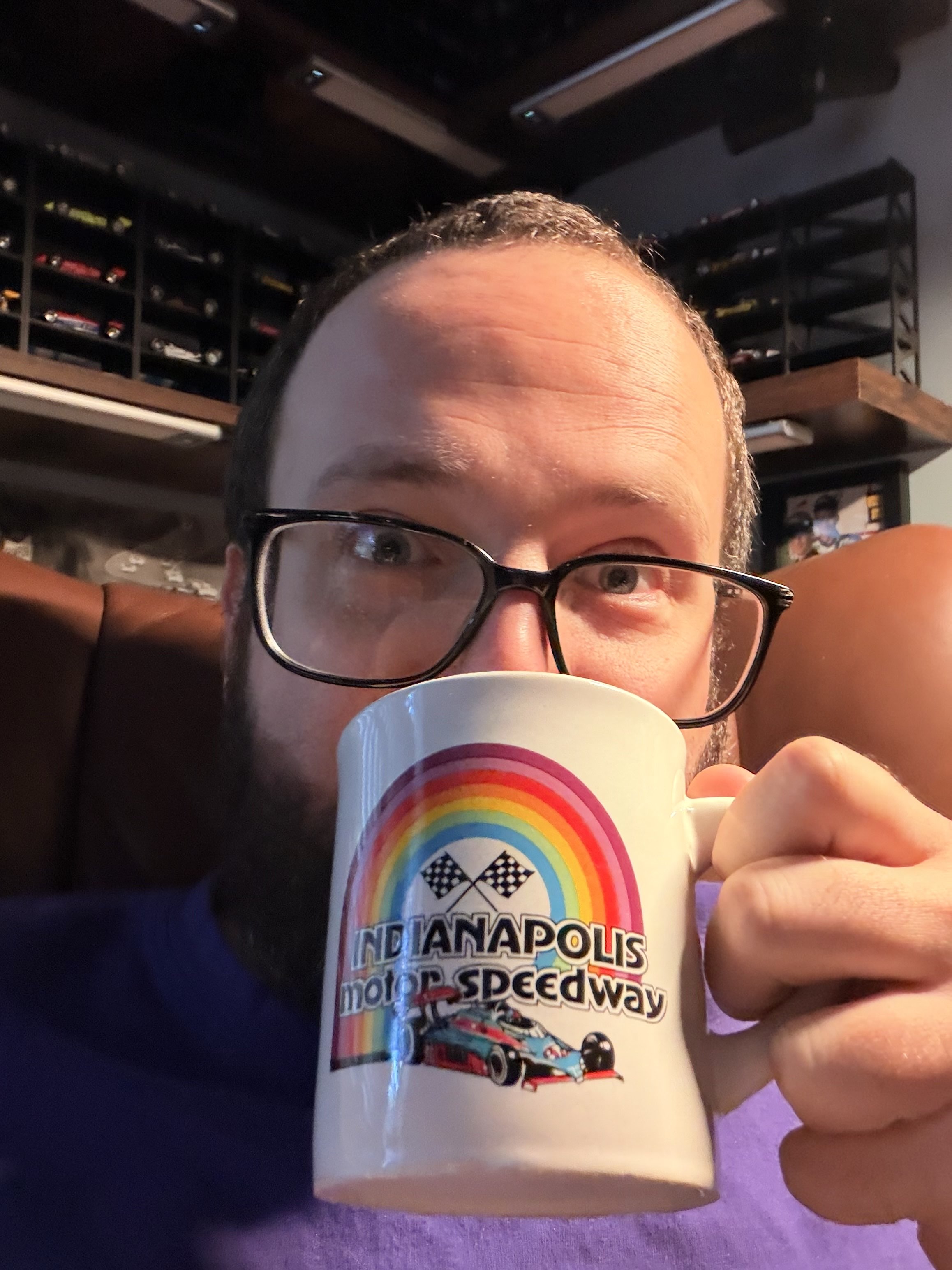 A man in glasses sipping coffee out of a ’70s-style Indianapolis Motor Speedway mug in a leather chair in front of shelves upon shelves of model race cars