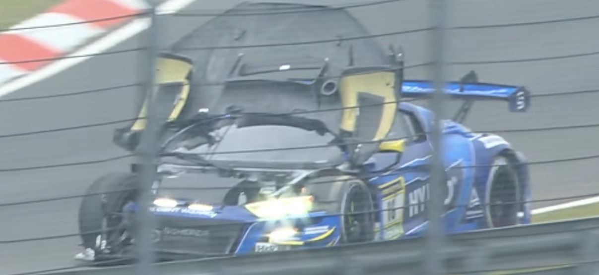 An Audi race car driving on the grass with its hood blown up onto its windshield