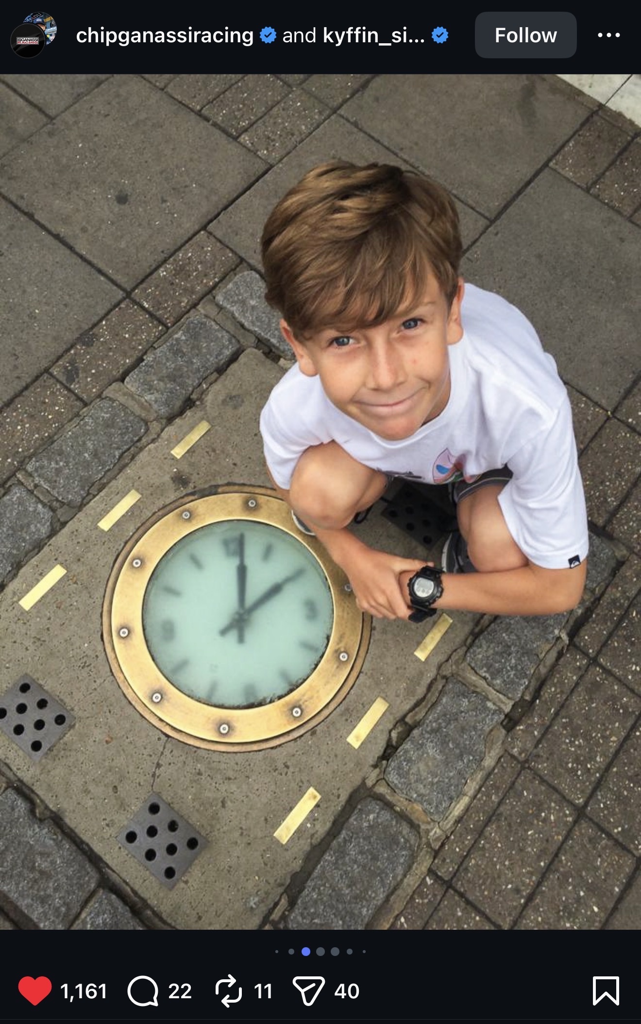 A young mop-headed Kyffin Simpson smiling wisely up from an ornate old-fashioned clock embedded into a sidewalk. He is wearing a cool watch.